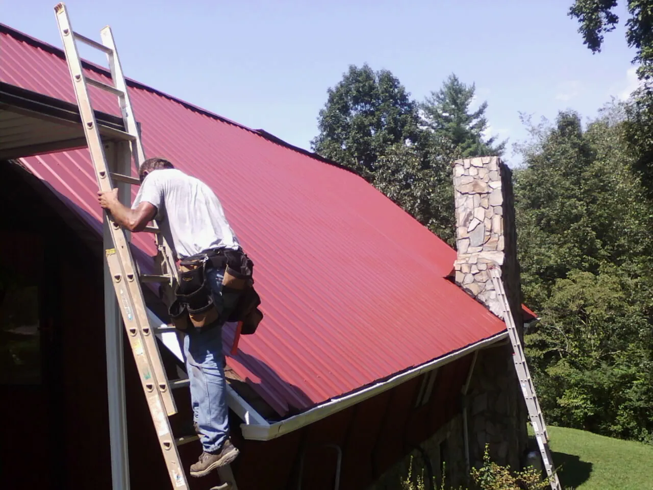 Roofer on a ladder beside a red metal roof and stone chimney — Soffit & Fascia Repair by Roofing Brothers in Albert Lea